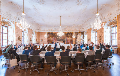 Versammlung in der steirischen Landstube: Teilnehmende sitzen an einem Tisch, viele heben die Hand (Abstimmung).